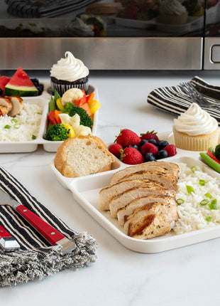 Nordic Ware divided meal trays with chicken rice vegetables fruit and cupcakes near microwave