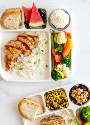Two Nordic Ware divided meal trays filled with colorful food portions including chicken rice vegetables fruit and dessert